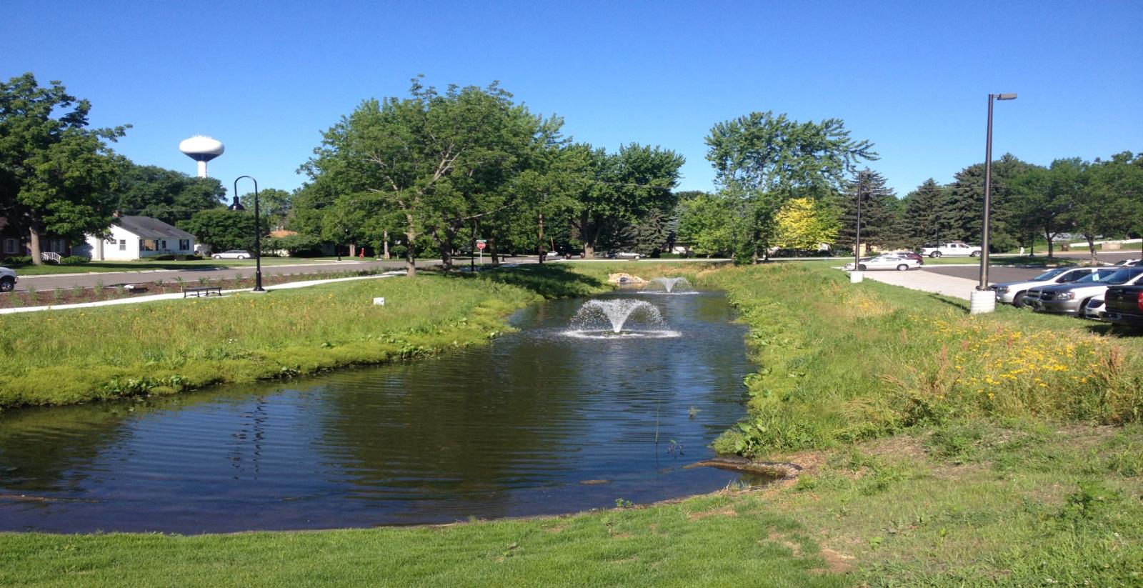 A narrow long pond with fountains and grassy banks on either side, in a city